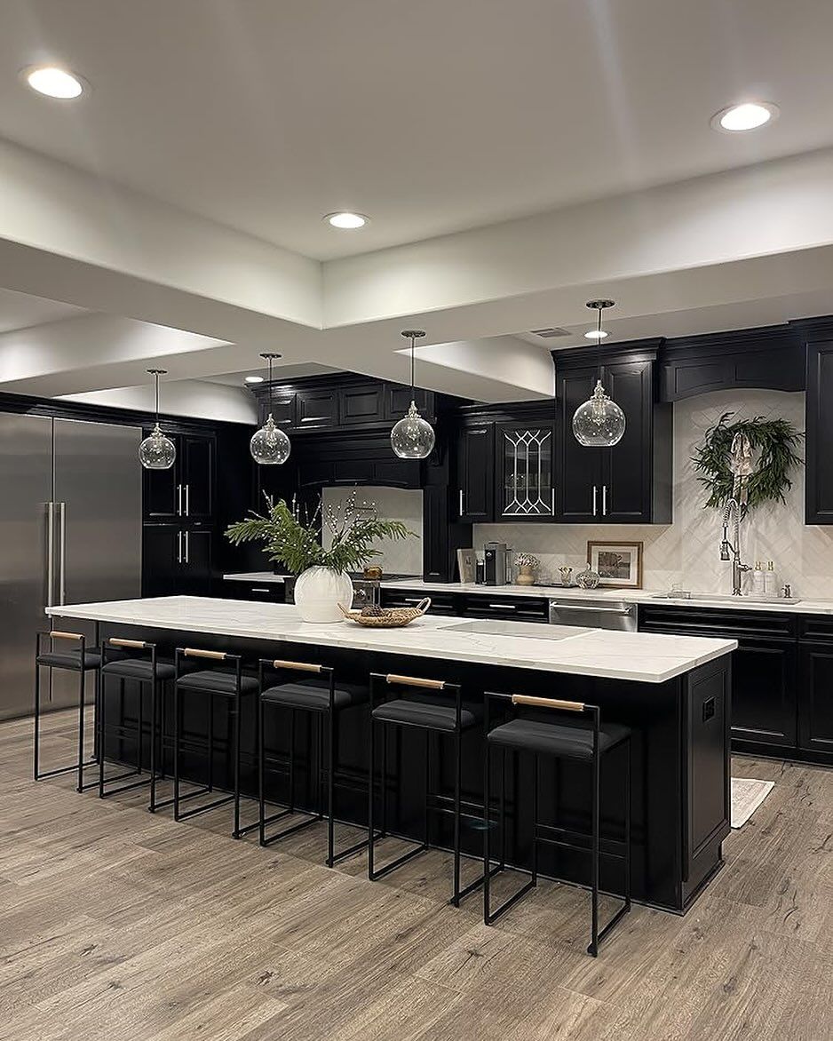 Modern kitchen with a long white-topped island, dark cabinetry, stainless steel appliances, and hanging glass pendants.