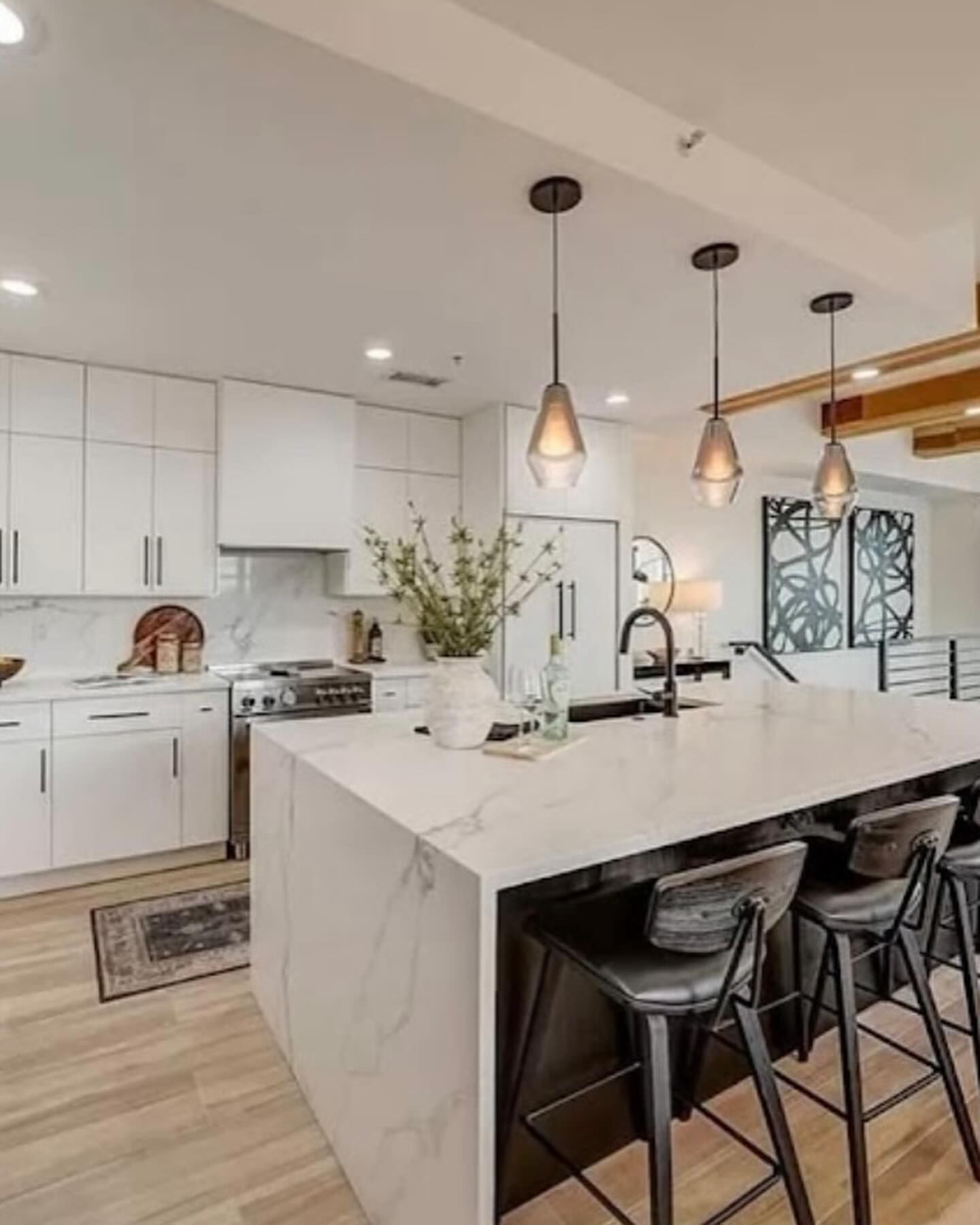 A modern white kitchen featuring a marble-topped island with three bar stools, pendant lighting, and wooden flooring.