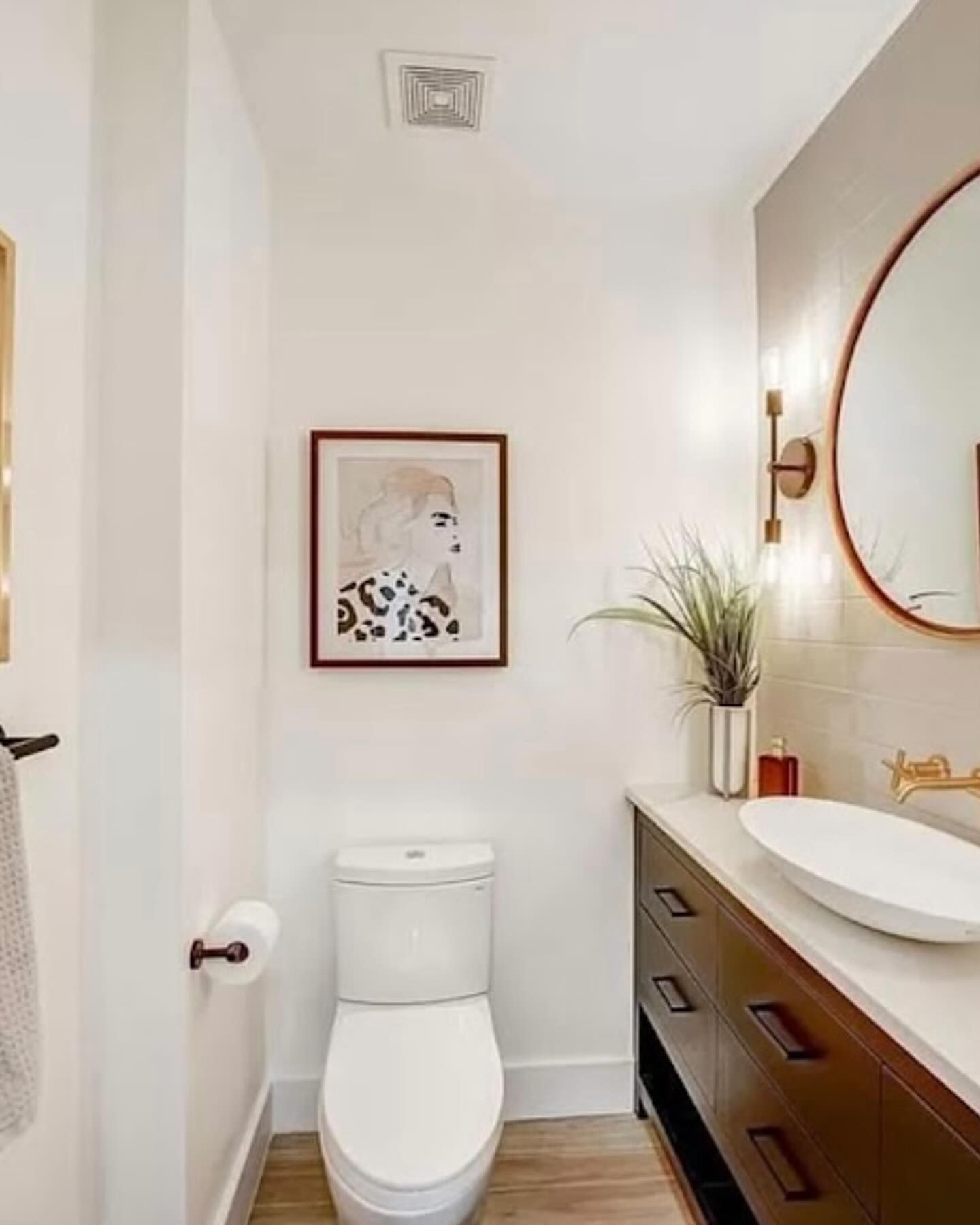 A modern bathroom with a white toilet, brown vanity, vessel sink, round mirror, and framed portrait art on the wall.
