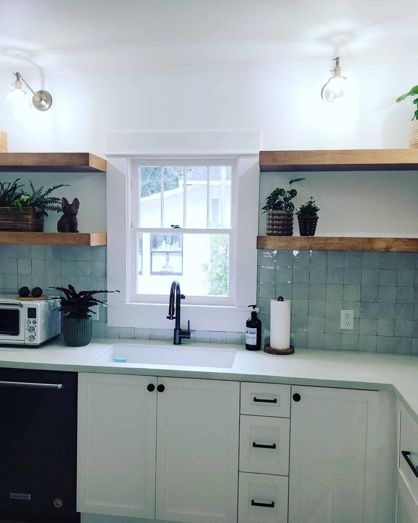 A kitchen featuring white cabinets, a farmhouse sink, light blue tiled backsplash, and wooden floating shelves.