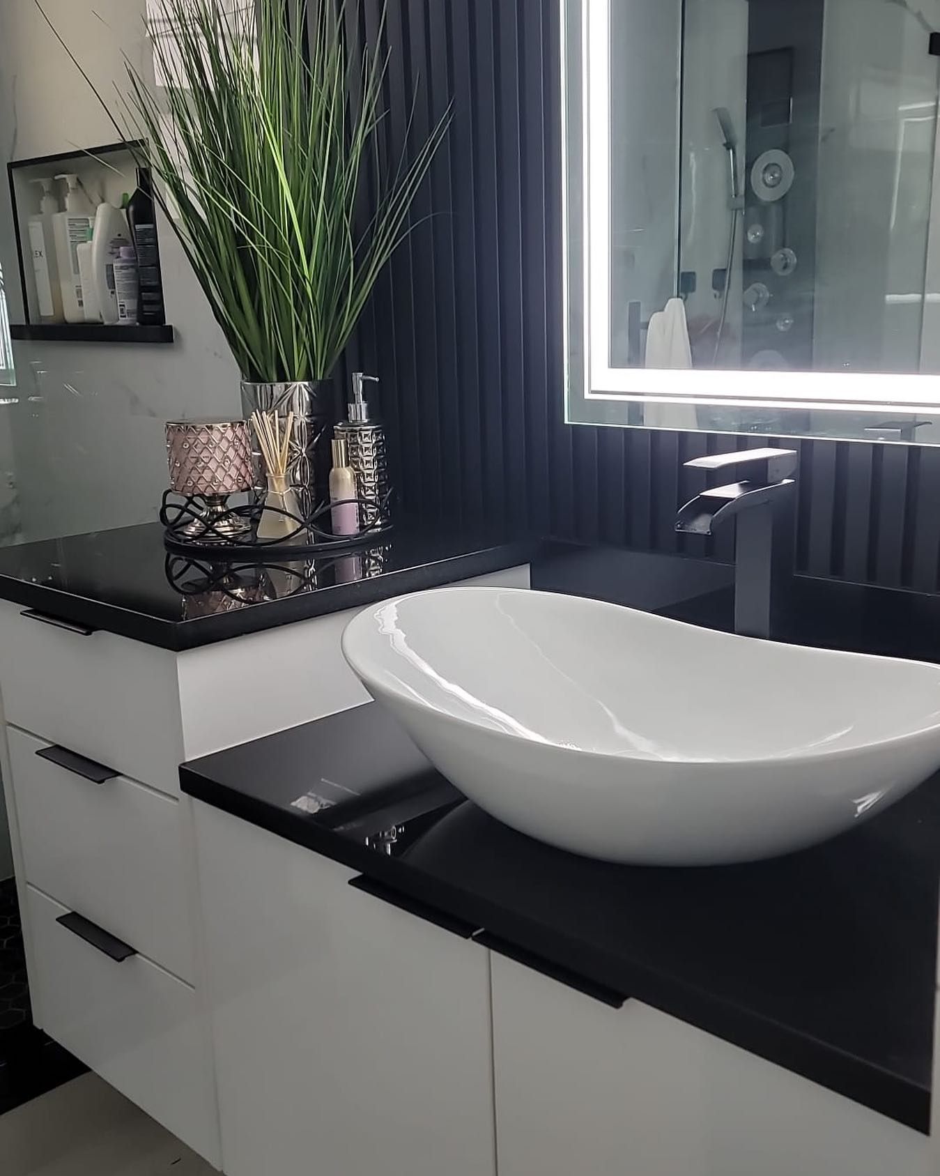 A modern bathroom vanity with a white vessel sink, black countertops, white cabinets, and a tall green plant decoration.