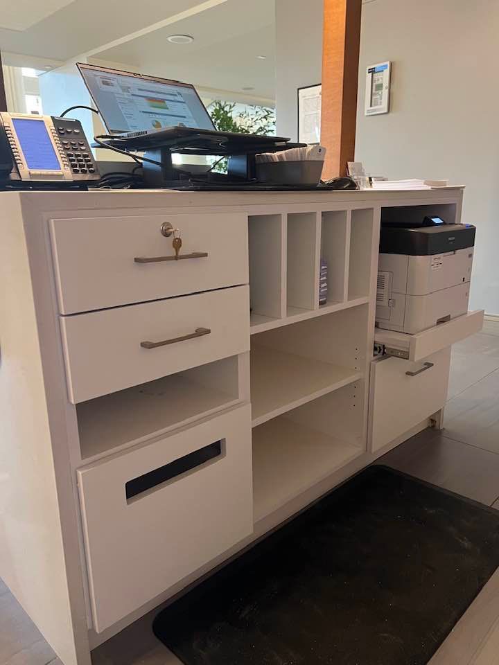 A white office reception desk with a computer, telephone, and printer in a bright, modern setting.