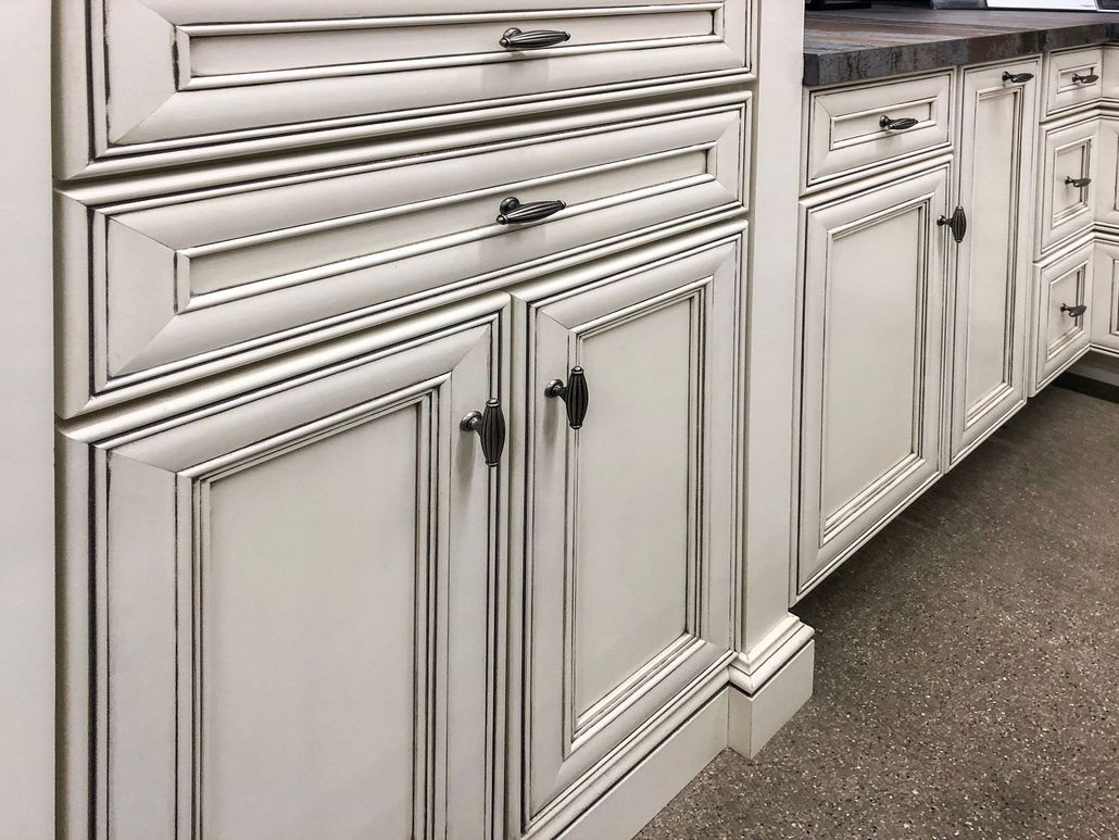 Off-white kitchen cabinets with a distressed finish, decorative molding, and dark hardware on a stone floor.