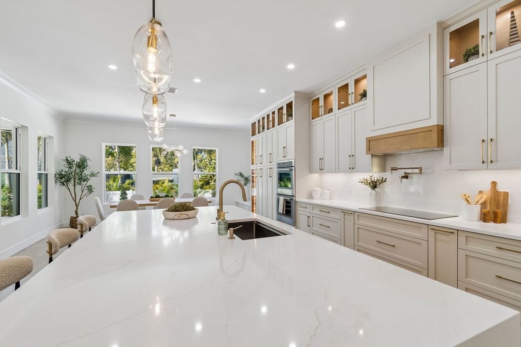 Bright modern white kitchen with island, brass pendant lights, and large windows overlooking greenery.