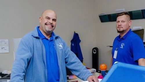 Two men in blue shirts smile at the camera. They stand in an office setting.