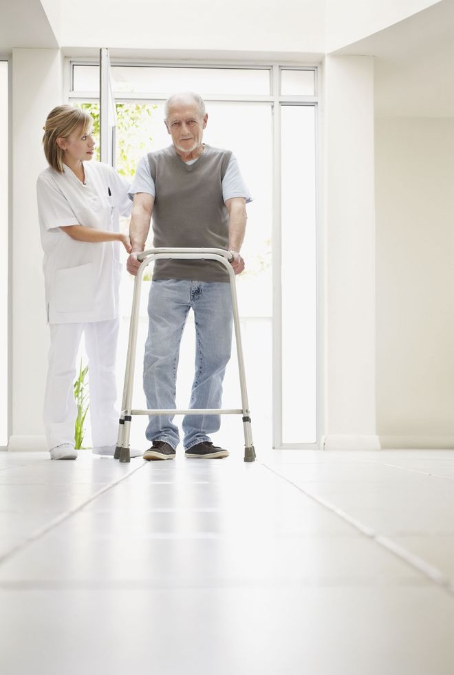 Nurse assisting a person using a walker in a brightly lit hallway.