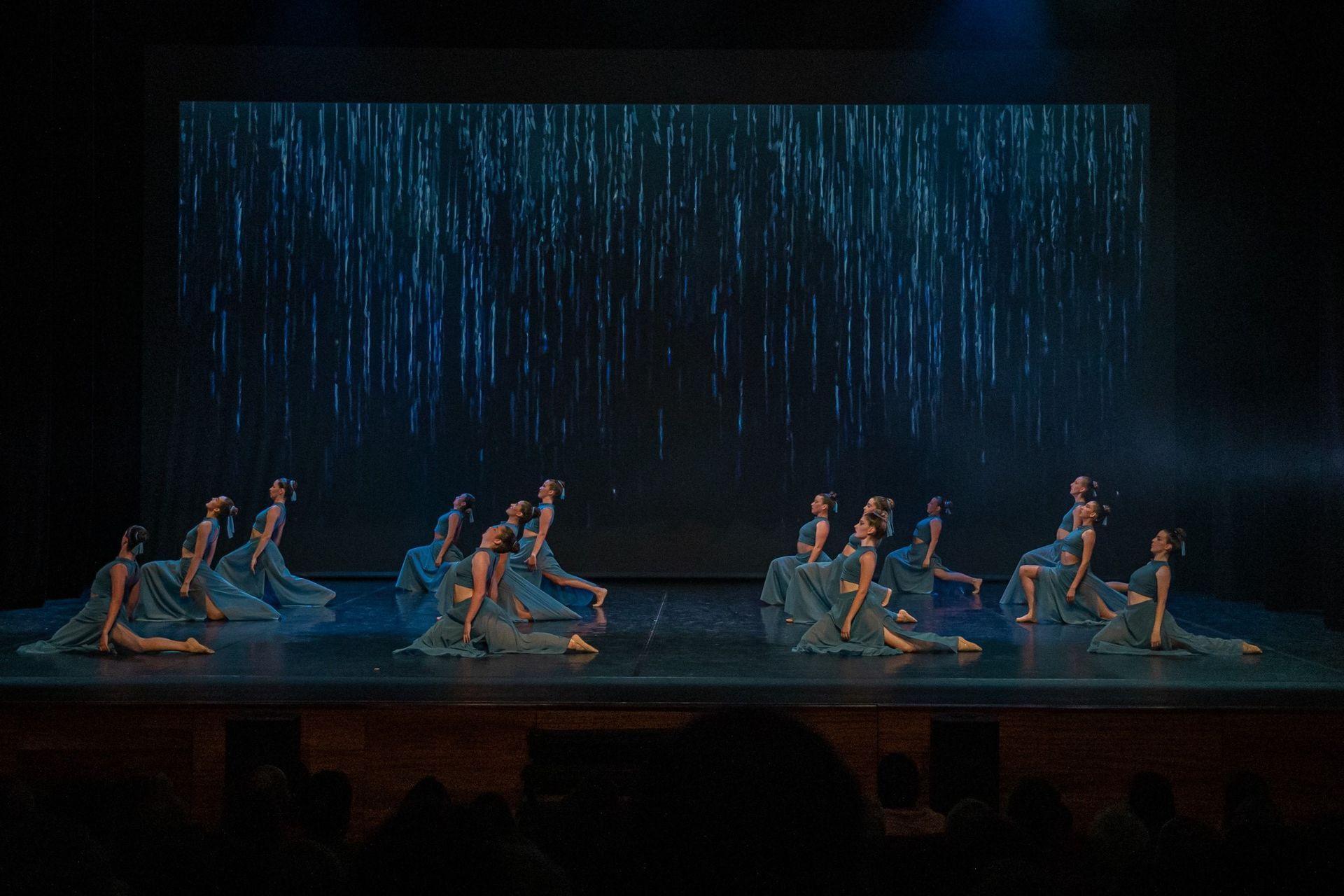 A group of dancers in teal dresses perform on a stage with a blue, rain-like backdrop. They are kneeling and reaching.