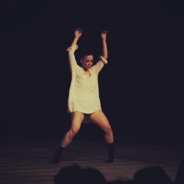 A woman with arms raised, in a white shirt and boots, dances on stage with a dark background.