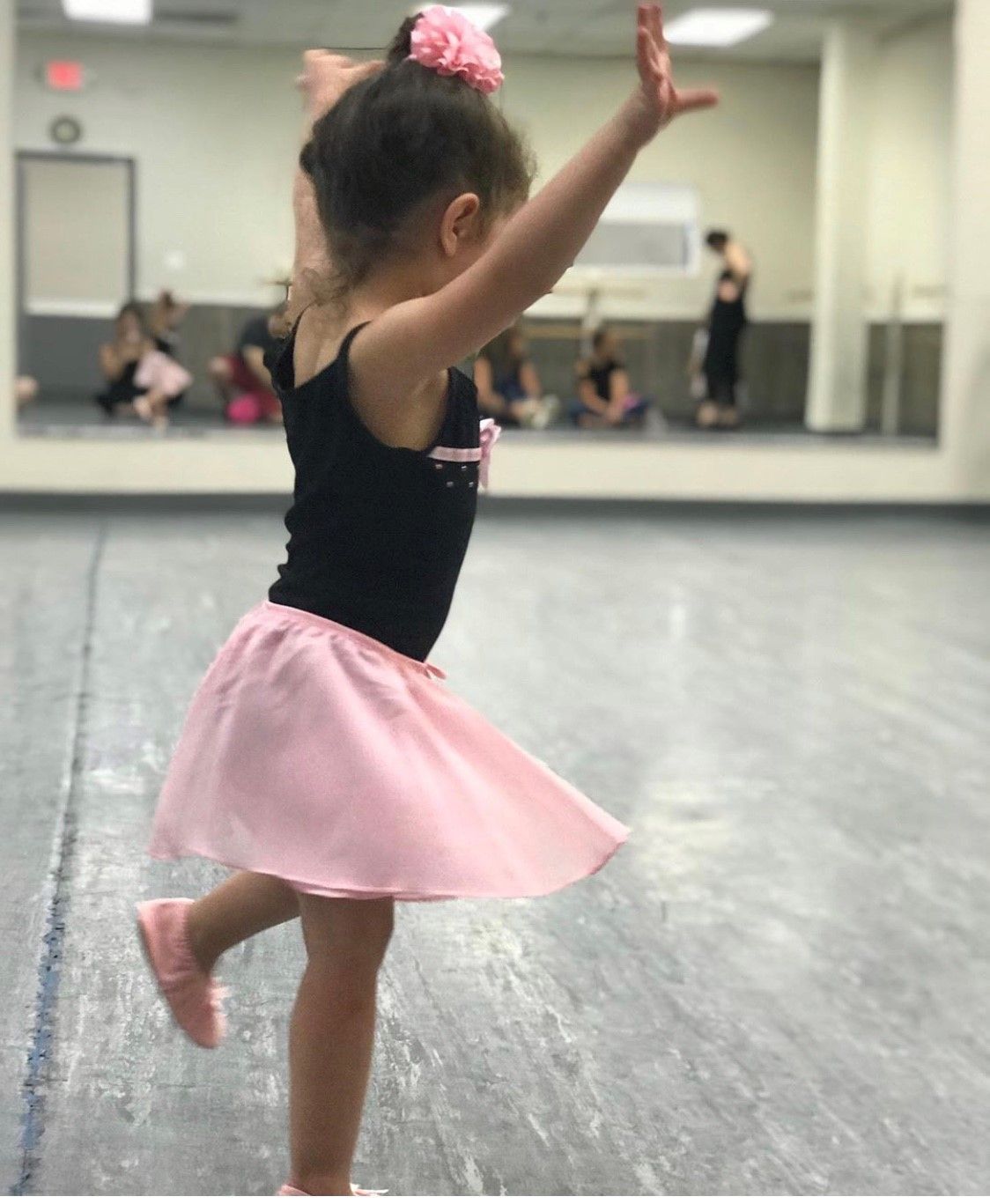 Young girl in a pink skirt and black leotard dances in a studio. She has her arms raised, and a pink flower in her hair.