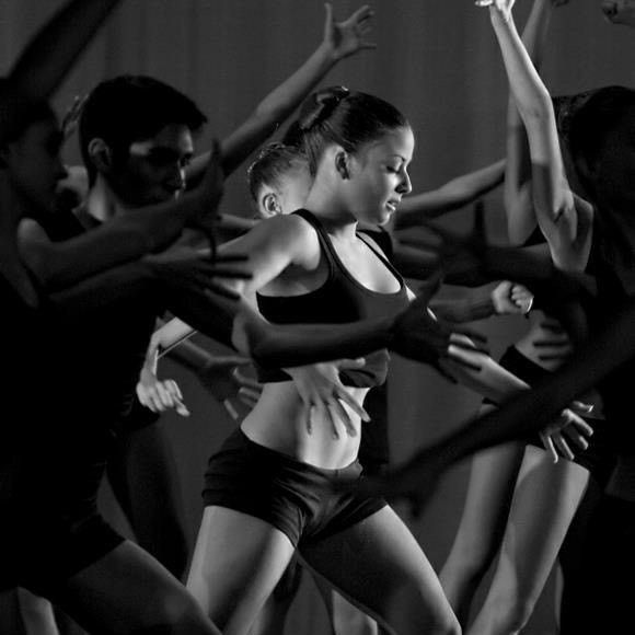 Black and white dance performance. A woman in a sports bra and shorts is center stage, surrounded by dancers reaching out.
