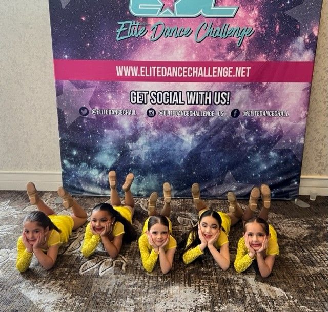 Five young girls in yellow outfits lie on their stomachs, smiling.