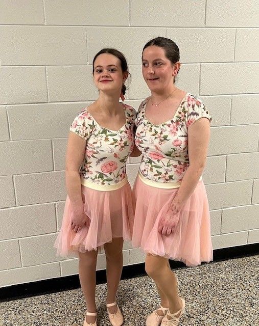 Two women in matching floral leotards and pink tutus stand against a brick wall, smiling. They wear ballet shoes.