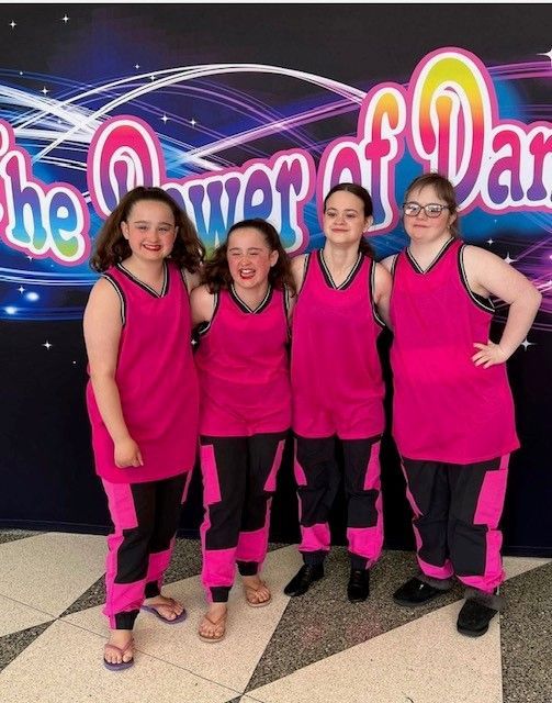 Four young women in matching pink and black dance outfits, posing in front of a