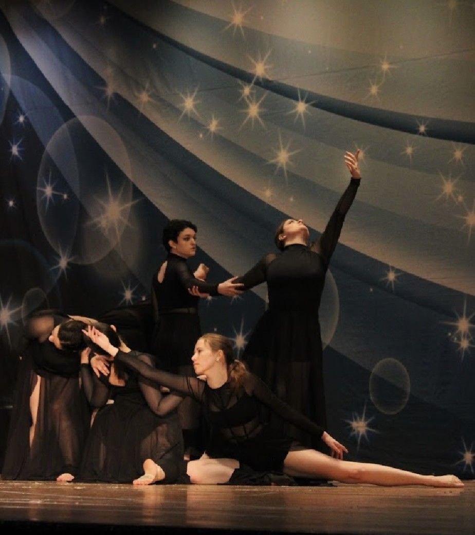 Dancers in black performing on a stage with a blue backdrop, one arm raised, others with arms outstretched.