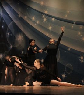 Dancers in black performing on a stage with a blue backdrop, one arm raised, others with arms outstretched.