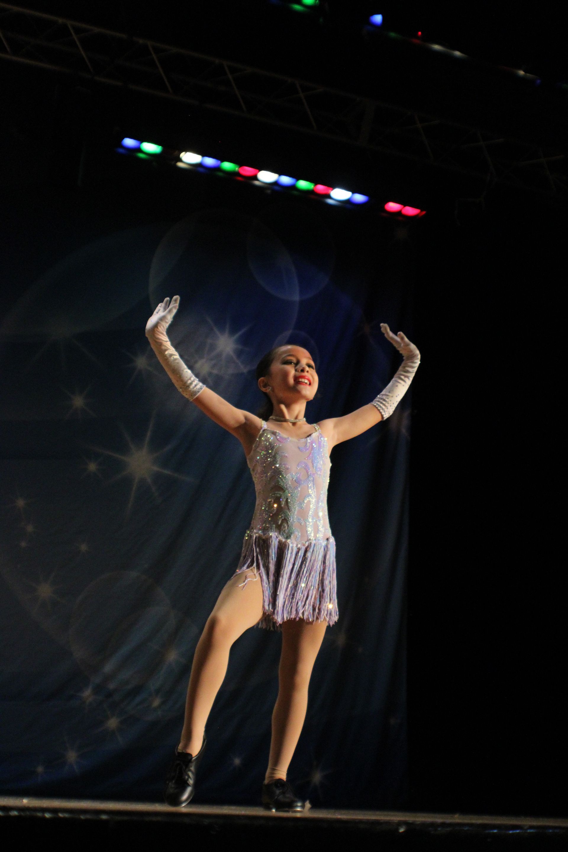 A young dancer on stage in a sparkly costume, arms raised, with stage lighting.