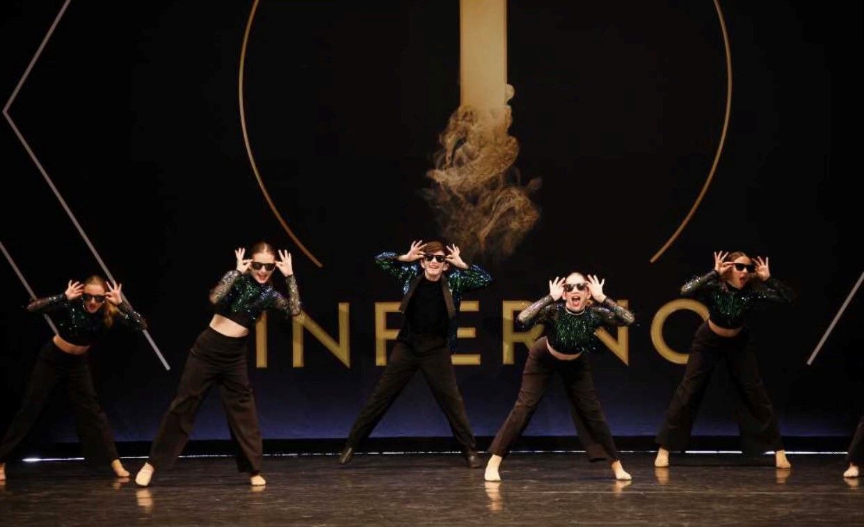 Five dancers in matching outfits perform on stage. They wear black pants, crop tops, and sunglasses, with hands framing their faces. The backdrop reads “Inferno”.