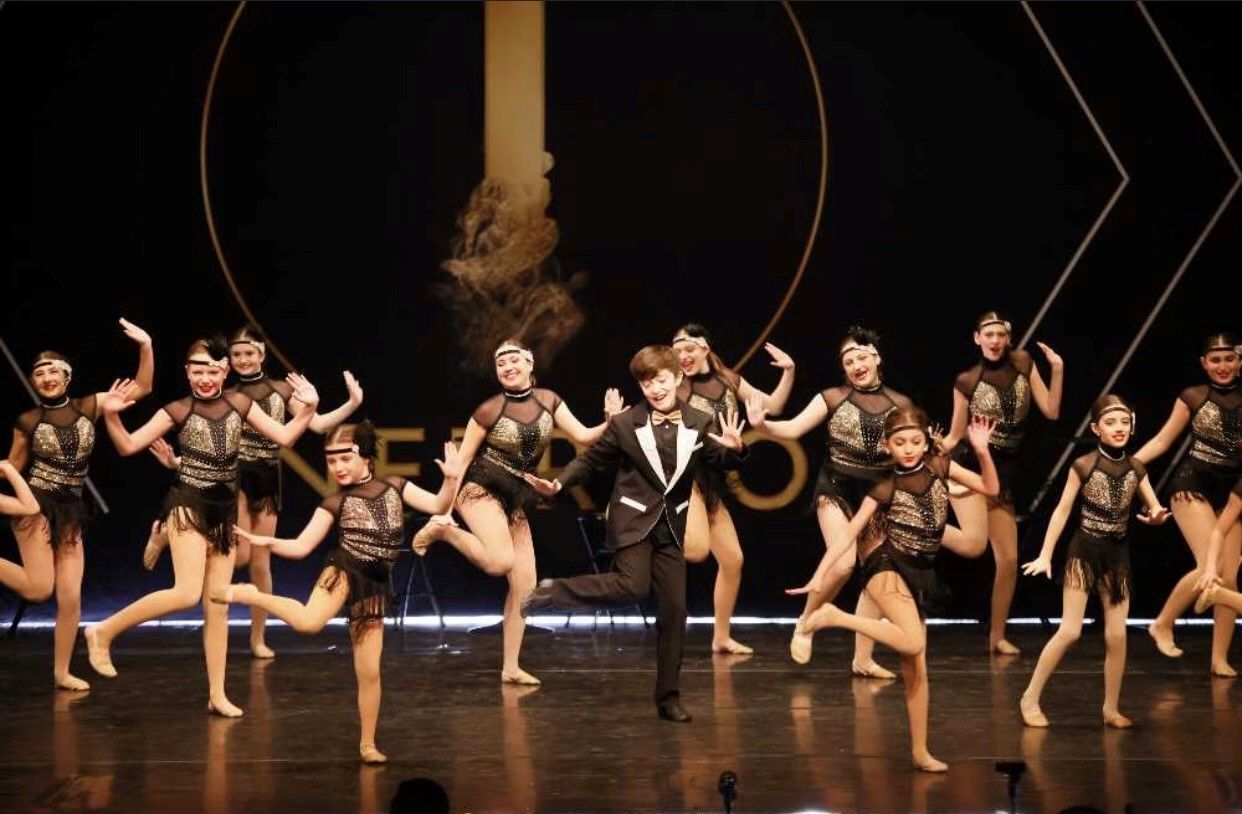 A dance ensemble in a stage production, dressed in black fringed outfits. A man in a tuxedo dances in the center. Gold details are visible.