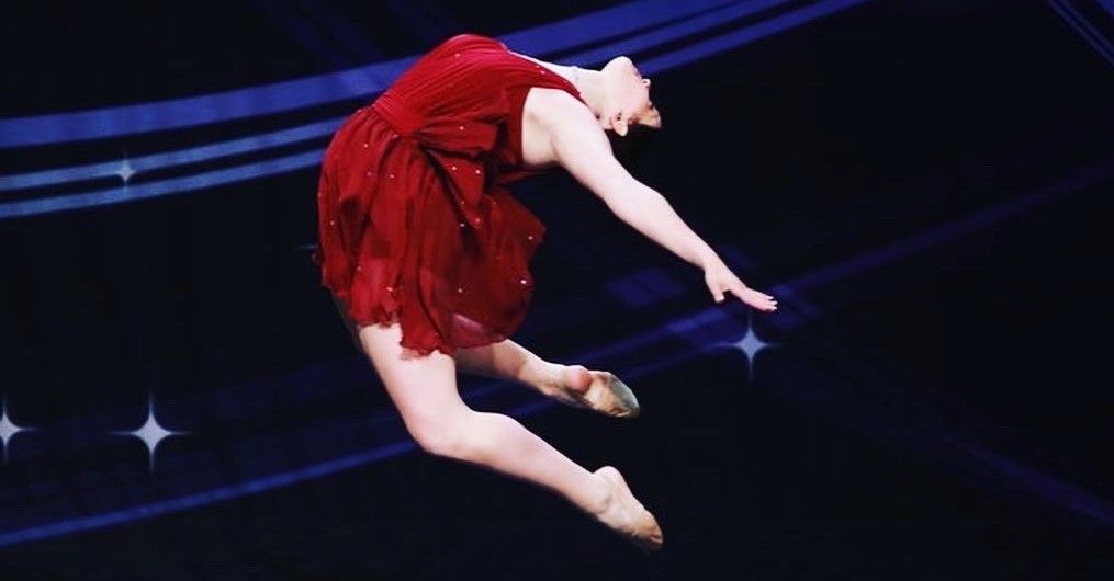 A dancer in a red dress leaps in the air against a dark background with star accents, performing a ballet move.