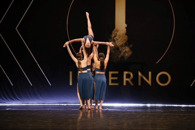 Dancers in teal costumes perform a lift on a stage with the word
