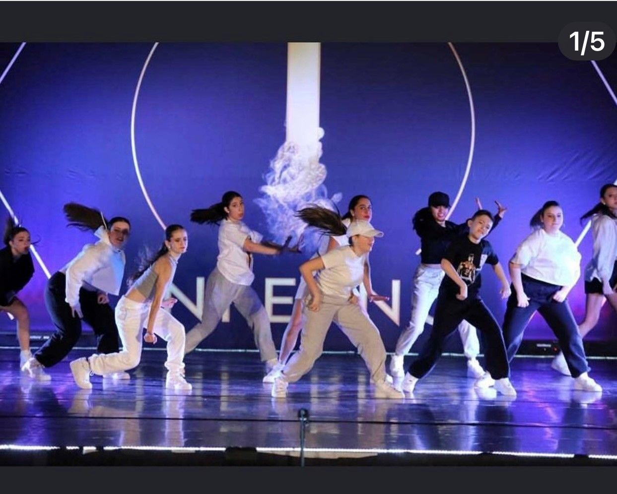 A group of dancers perform on stage. They wear casual attire and move in unison against a blue backdrop.