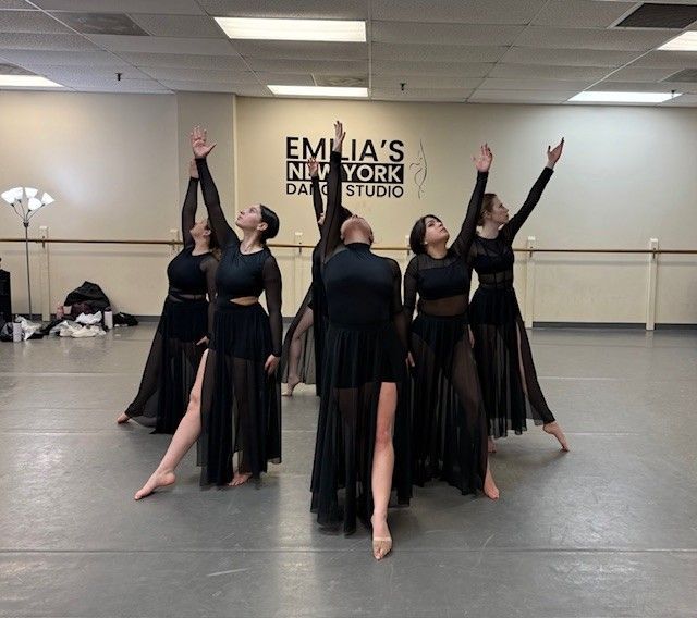 A group of dancers in black dresses with sheer skirts practice in a studio. They have their arms raised.
