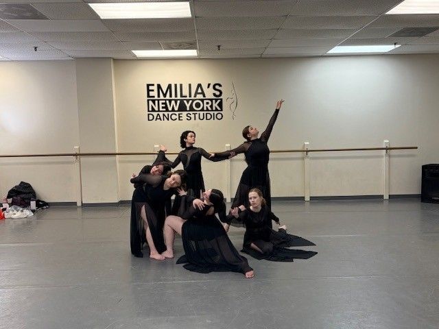 Dancers in black outfits perform in a dance studio. They are posed, some reaching up with arms, others kneeling.