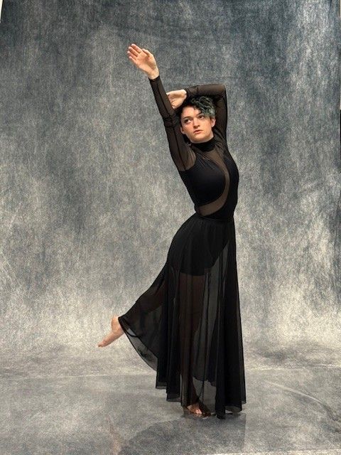A dancer in a black dress with sheer skirt poses on a gray background. One arm is raised overhead, other bent at the head.