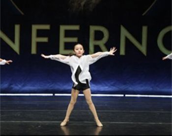 A young girl dances on a stage, arms outstretched. She wears a white shirt over a black outfit
