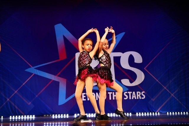 Two young dancers in black and red costumes performing on stage in front of a blue backdrop with a star logo.