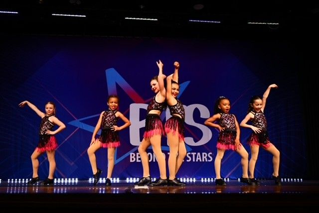 A dance troupe of six young girls in black and red outfits performs on a stage.