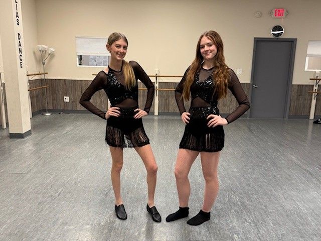 Two young women in black dance costumes pose in a dance studio. They have long hair and are smiling, with hands on their hips.