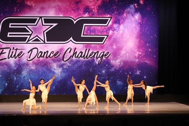 A dance troupe in cream-colored costumes performs on stage, with a galaxy backdrop