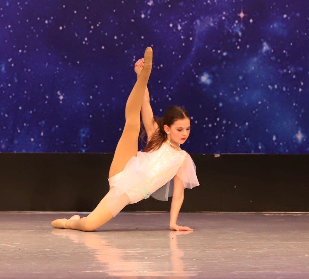 A young dancer in a white leotard performs a split with an elevated leg, set against a starry backdrop.