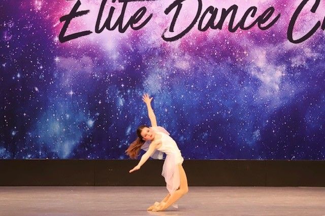 Dancer in a white dress performing on stage in front of a starry backdrop; bent backward with arms raised.