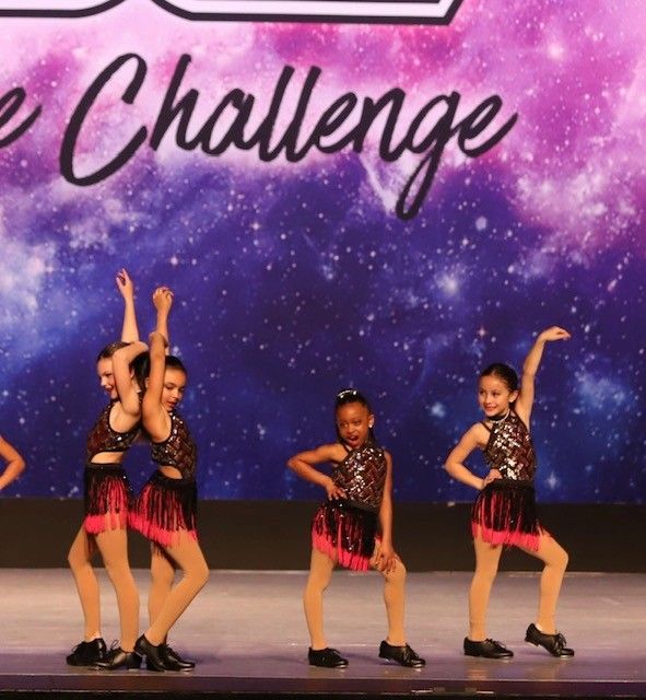 Young dancers in matching costumes perform on a stage with a galaxy backdrop. They strike poses with arms raised.