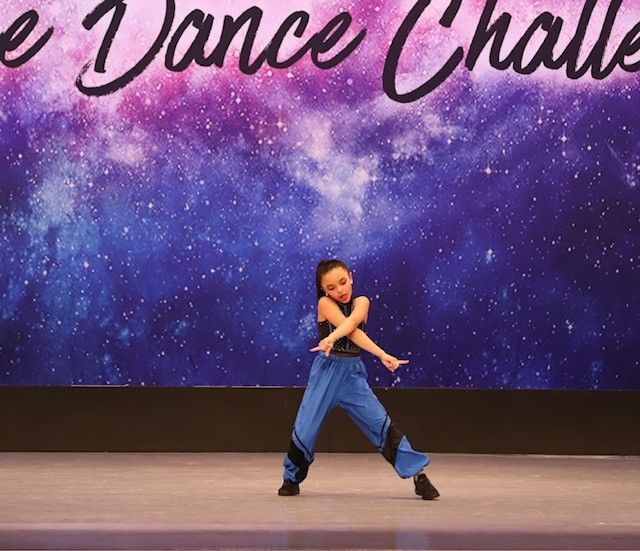 Young dancer performing on stage in front of a starry backdrop; she wears blue pants, black top, and has her arms crossed.
