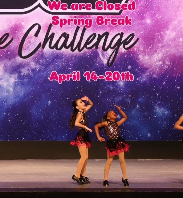 Two young dancers in black and red costumes perform on stage, with a cosmic background