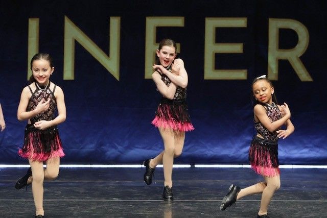 Three young girls tap dancing on stage, wearing black and pink fringed costumes.