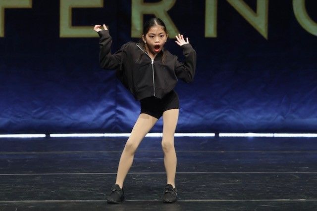 Young dancer on stage, arms raised, mouth open in surprise. She wears a black jacket, shorts, and tights.
