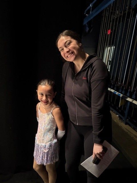 A young girl in a sparkly ice skating dress smiles next to a woman in black, holding papers, backstage in a dark theater setting.