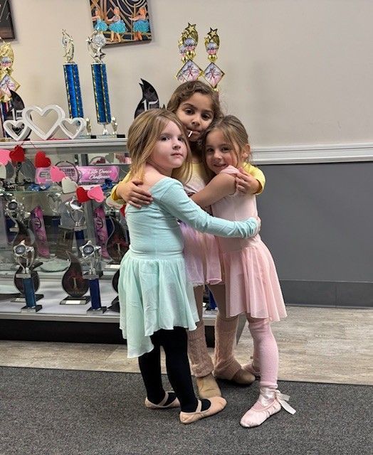 Three young girls in ballet attire embrace, smiling. They are standing in a dance studio.