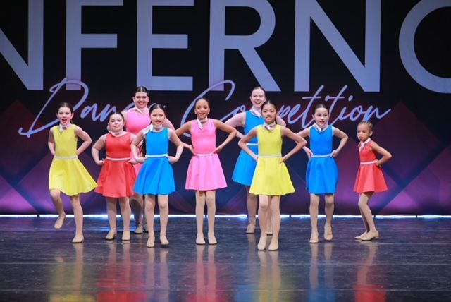 Seven dancers in bright costumes pose on a stage with an “INFERNO” backdrop