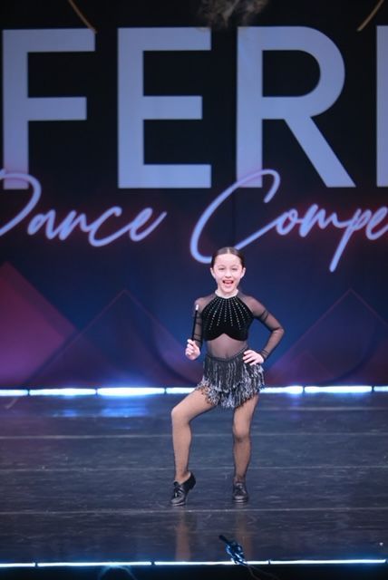 Young dancer in black costume performing on stage with a purple “dance competition” backdrop