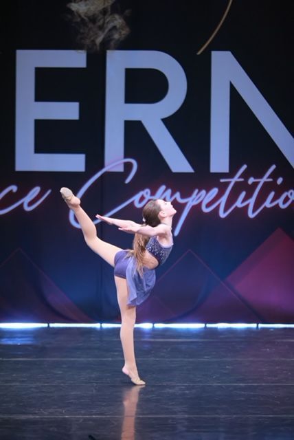 Young dancer in a blue costume balancing on one leg on a competition stage under purple lighting