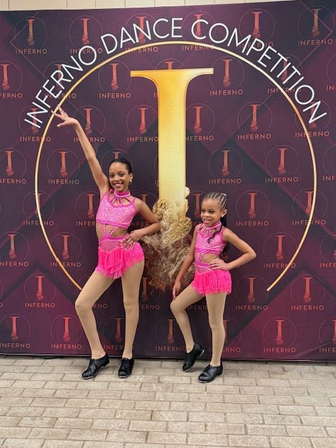 Two young dancers in pink costumes posing in front of an Inferno Dance Competition backdrop.