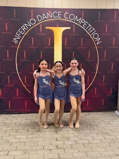 Three dancers in blue costumes posing in front of an Inferno Dance Competition backdrop
