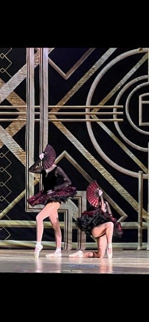 Two ballerinas in black and burgundy costumes perform on stage with an art deco backdrop. One is standing en pointe, and the other is kneeling.
