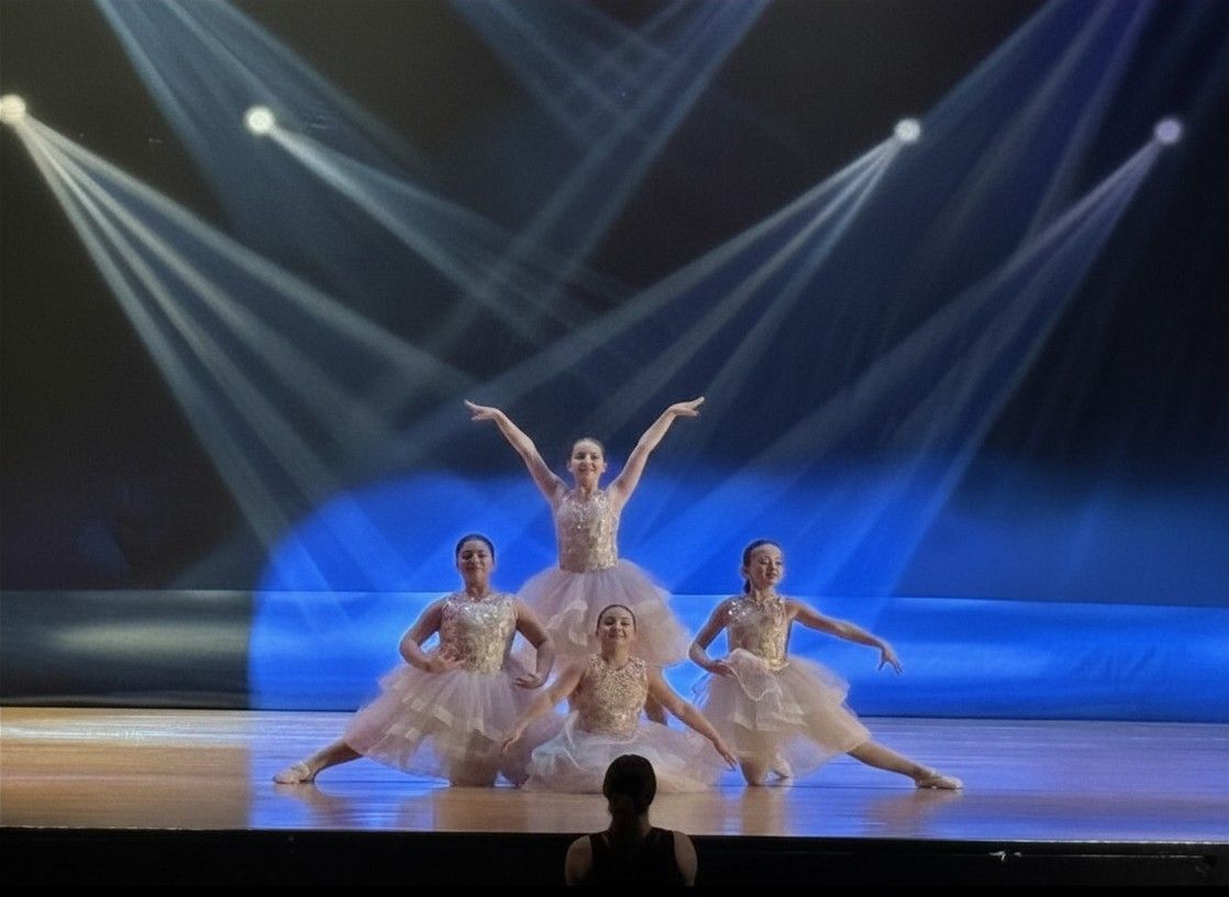 Three dancers in white costumes pose on a lit stage with blue spotlights overhead.
