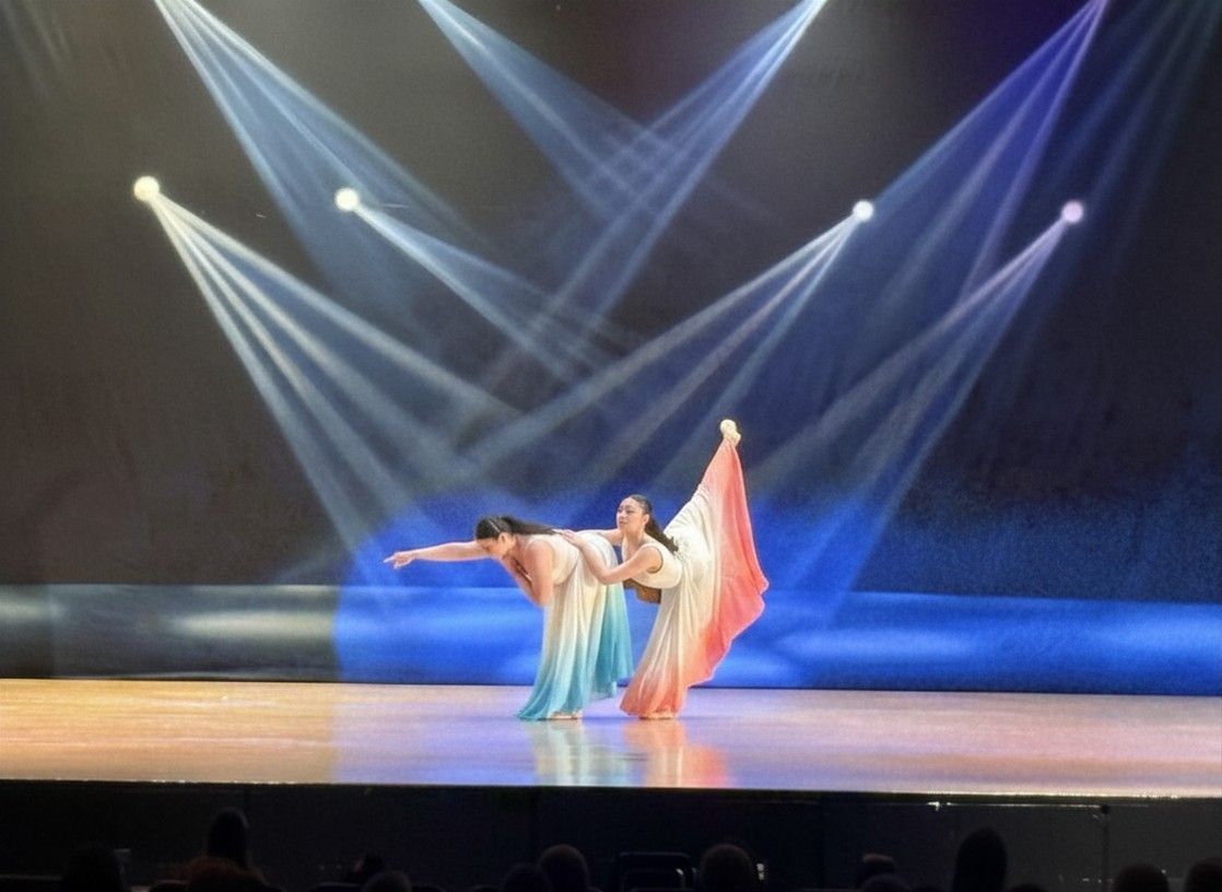 Two dancers in flowing pastel costumes perform under blue stage spotlights.